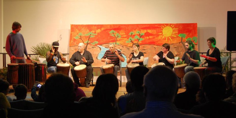 Concert de percussion avec plusieurs musiciens sur scène, devant un public attentif. Décor coloré en arrière-plan.