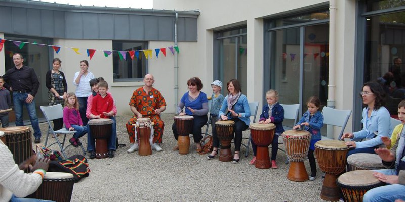 Atelier de percussion en plein air avec des participants de tous âges, devant un bâtiment de la commune.