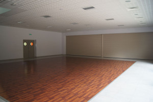 Intérieur d'une salle polyvalente à Feuguerolles-Bully, sol en parquet, murs lumineux et rideaux métalliques fermés.