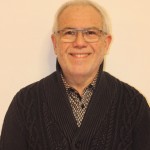 Portrait d'un homme souriant portant un pull noir, sur fond clair.