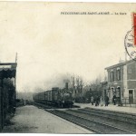 Gare de Feuguerolles-Saint-André avec train à vapeur et passagers sur le quai, époque vintage.