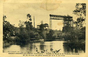 Vue d'une ancienne installation industrielle à Saint-André-sur-Orne, près d'un plan d'eau et entourée de végétation.