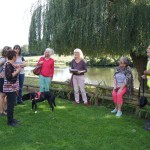 Groupe de personnes réunies au bord d'un plan d'eau, avec un chien, sous un arbre, par une journée ensoleillée.