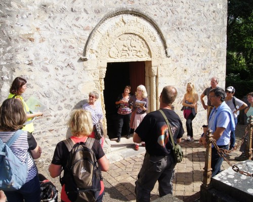 Visite guidée d'un groupe devant une église à Feuguerolles-Bully, avec une guide présentant des informations sur le site.