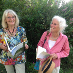 Deux femmes souriantes tiennent des livres en pleine nature, entourées de verdure et de fleurs.