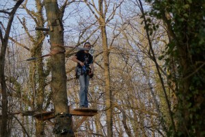 Homme sur une plateforme en bois dans un arbre, équipé pour l'escalade, entouré d'une forêt.