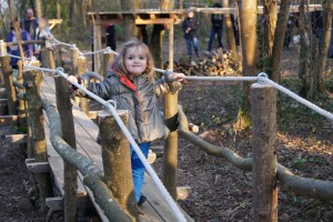 Une petite fille souriante traverse un pont en bois, entourée par des arbres et des adultes au second plan.