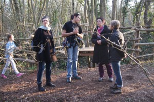 Groupe de personnes discutant en pleine nature, avec une enfant en arrière-plan, près d'une plateforme dans les arbres.