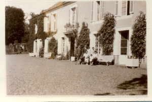 Photo en noir et blanc d'une famille posant devant une maison avec des volets, entourée de verdure.