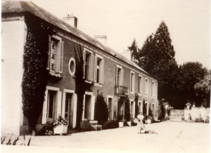 Ancienne maison avec des volets et des plantes grimpantes, animée par des personnes sur une cour ensoleillée.