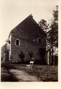 Maison en pierre avec un chemin, une chaise blanche et des chaises empilées dans un jardin. Photo en noir et blanc.
