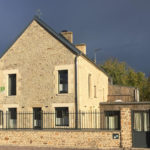 Maison rénovée en pierre située à Feuguerolles-Bully, avec une clôture et un mur en pierre. Ciel menaçant en arrière-plan.