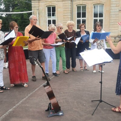 Groupe de choristes en répétition en plein air, avec un cadre rural et un bâtiment ancien en arrière-plan.