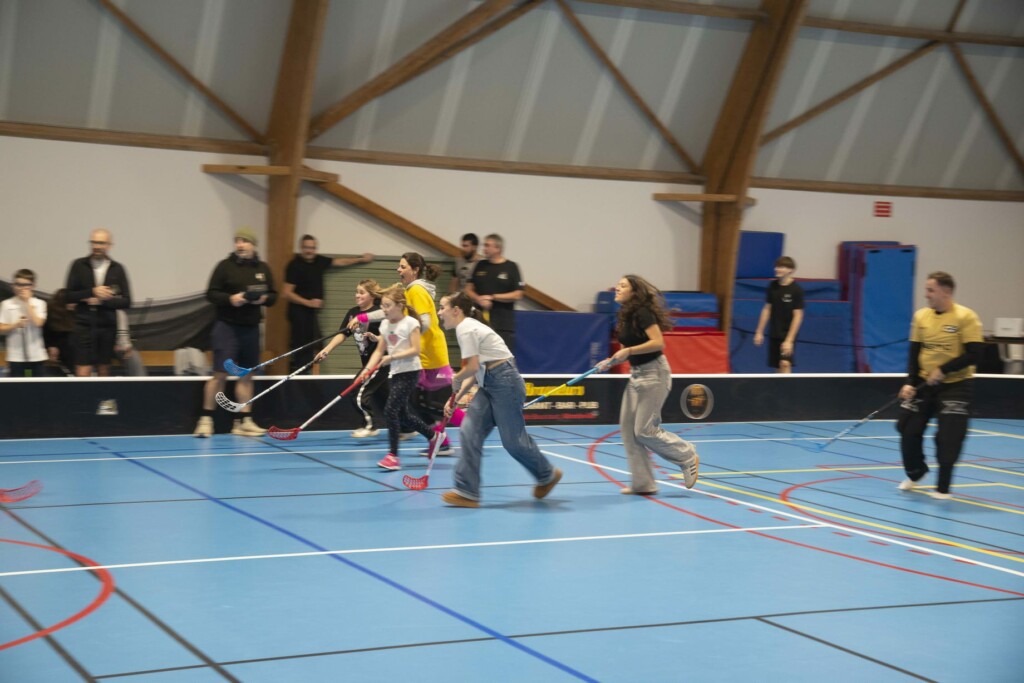 Jeunes joueurs de hockey en salle s'entraînant, avec des spectateurs en arrière-plan dans un gymnase.