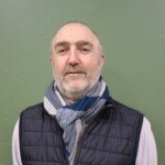 IMG_2526b Portrait d'un homme souriant portant une écharpe et un gilet, devant un mur vert.