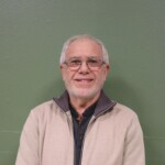 IMG_2527b Un homme souriant portant une veste beige, devant un mur vert.