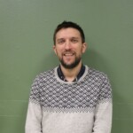 IMG_2529b Homme souriant portant un pull en laine, devant un mur vert, dans un cadre informel.