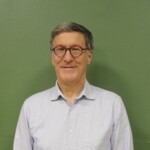 IMG_2530b Portrait d'un homme souriant, en chemise claire, devant un mur vert.