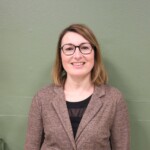 IMG_2531b Portrait d'une femme souriante, portant des lunettes et un blazer, devant un mur vert.