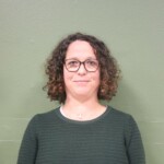 IMG_2533b Portrait d'une femme aux cheveux bouclés, portant des lunettes, sur fond vert.