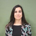 IMG_2538b Portrait d'une femme souriante, avec des cheveux longs et un vêtement à motifs floraux sur fond vert.