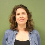 IMG_2544b Portrait d'une femme souriante, cheveux bouclés, portant un haut noir et une chemise en jean, devant un mur vert.