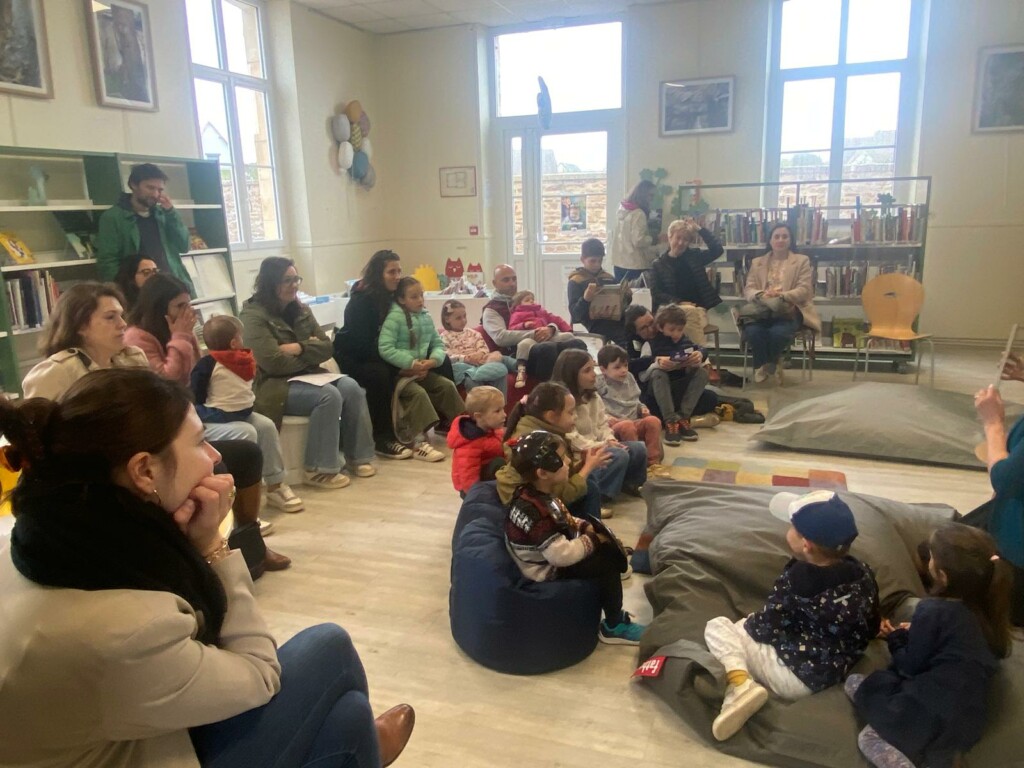 Atelier de lecture pour enfants, avec des adultes et des enfants assis sur des coussins dans une bibliothèque.