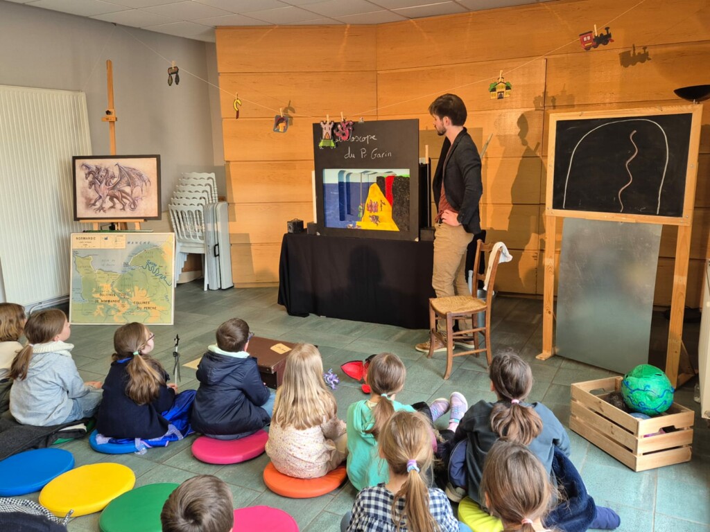 Atelier éducatif avec un intervenant et des enfants assis sur des coussins colorés, devant un tableau et une carte.