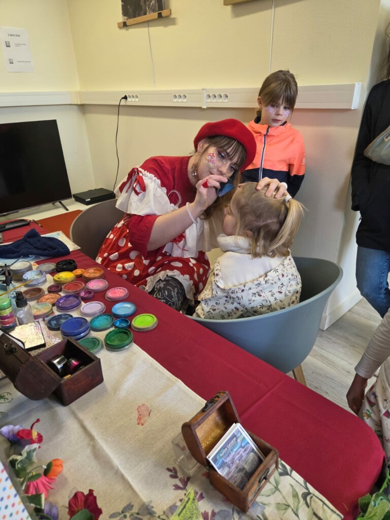 Une artiste maquille une petite fille pendant un événement convivial à Feuguerolles-Bully, avec des pots de peinture sur la table.