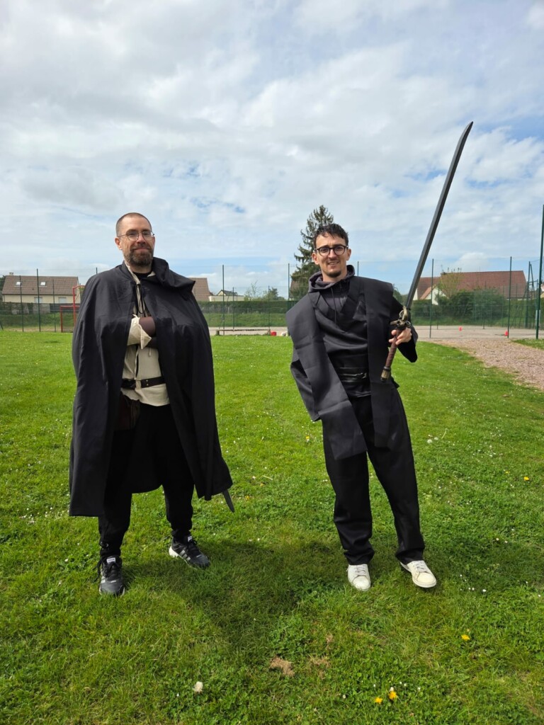 Deux hommes en costumes noirs, l'un avec une épée, posent sur un terrain vert sous un ciel nuageux.