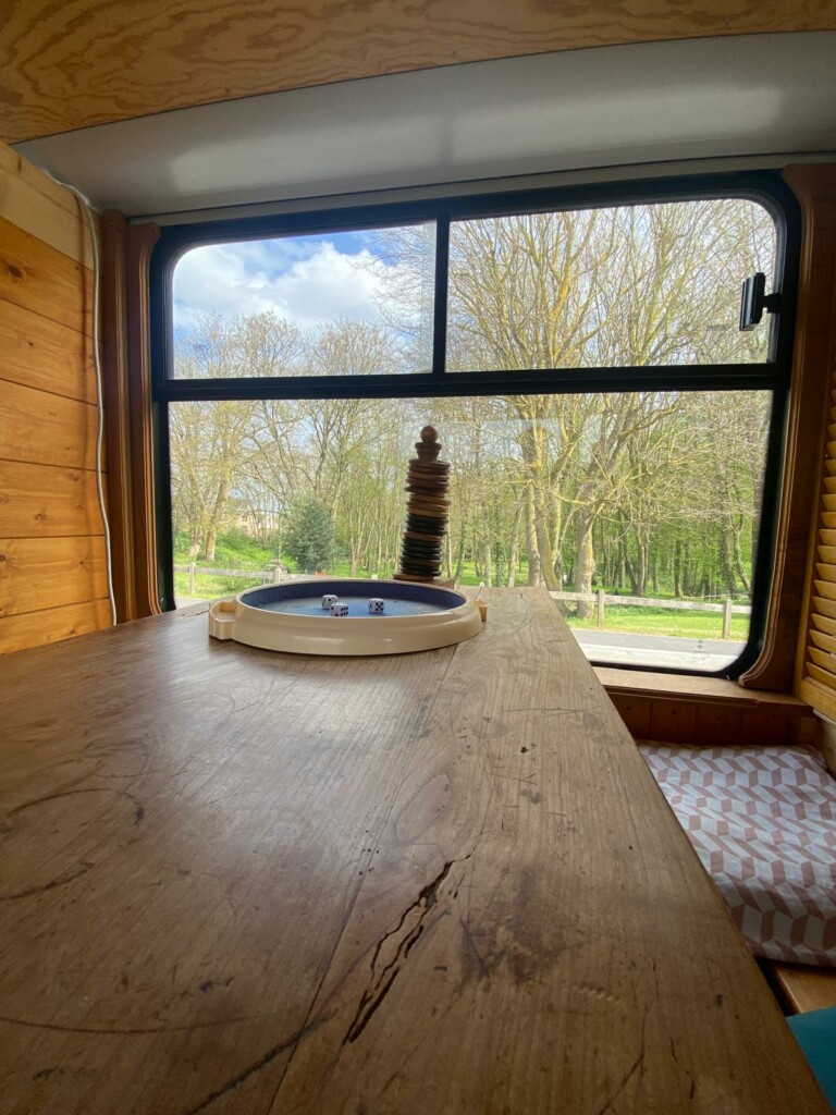 Une table en bois à l'intérieur d'un espace aménagé, avec des dés et une fenêtre donnant sur un paysage boisé.