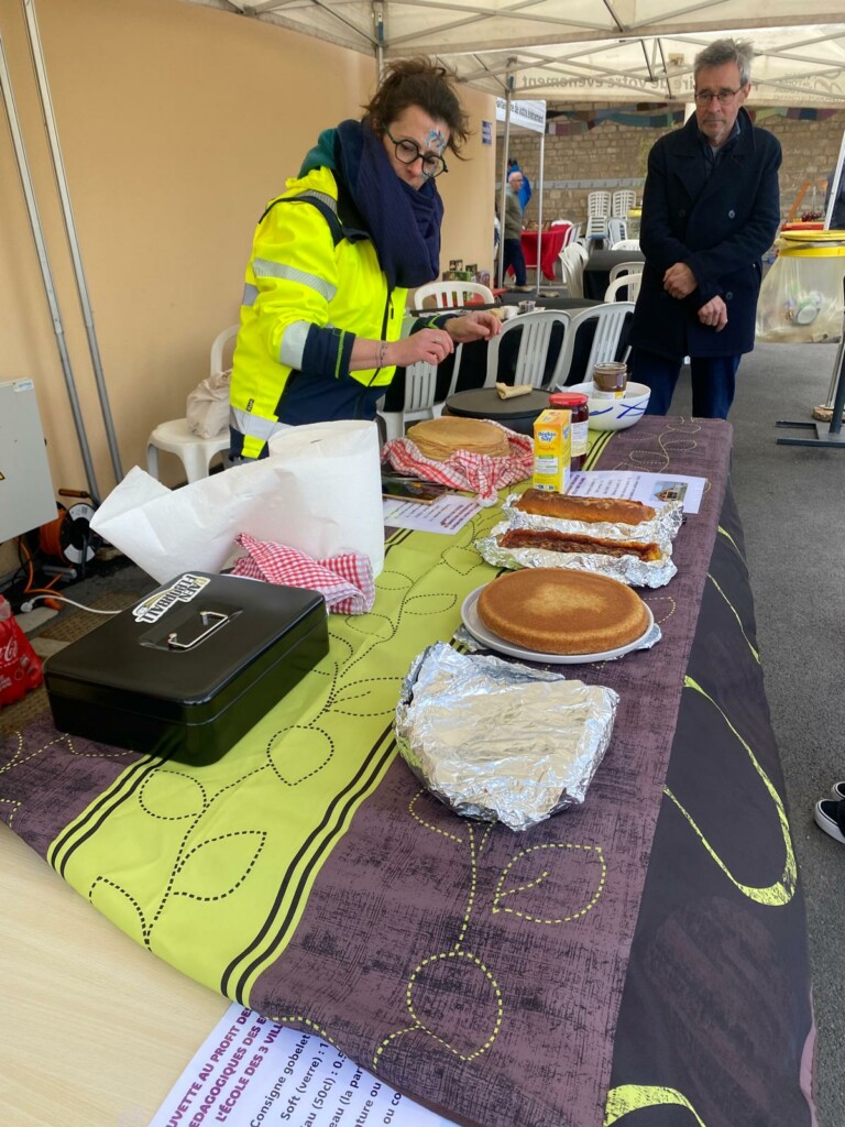 Femme en tenue de sécurité préparant des crêpes lors d'un événement communal, avec différents plats sur une table.