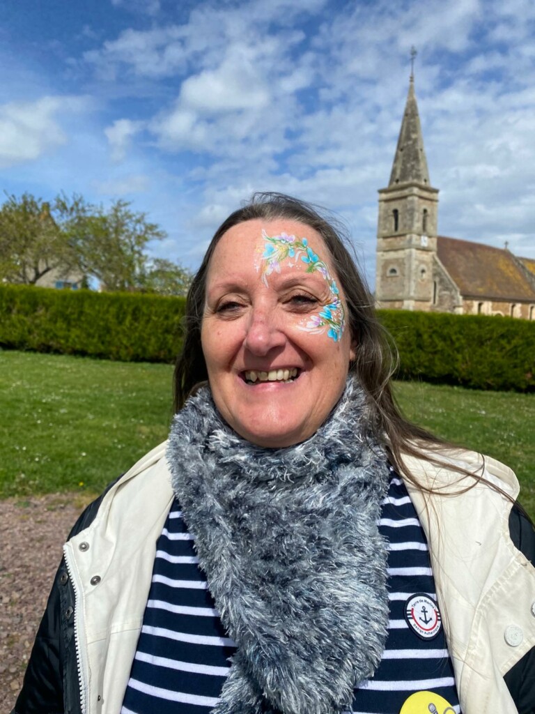 Femme souriante avec un visage peint, portant un écharpe grise, en arrière-plan une église et un ciel nuageux.