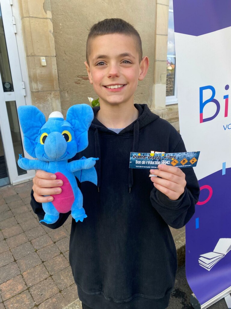 Un enfant souriant tient une peluche bleue en forme d'éléphant et un bon de réduction, devant un bâtiment municipal.