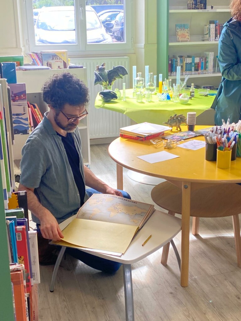 Un homme feuillette un livre sur les dragons dans une bibliothèque, entouré de matériel éducatif.