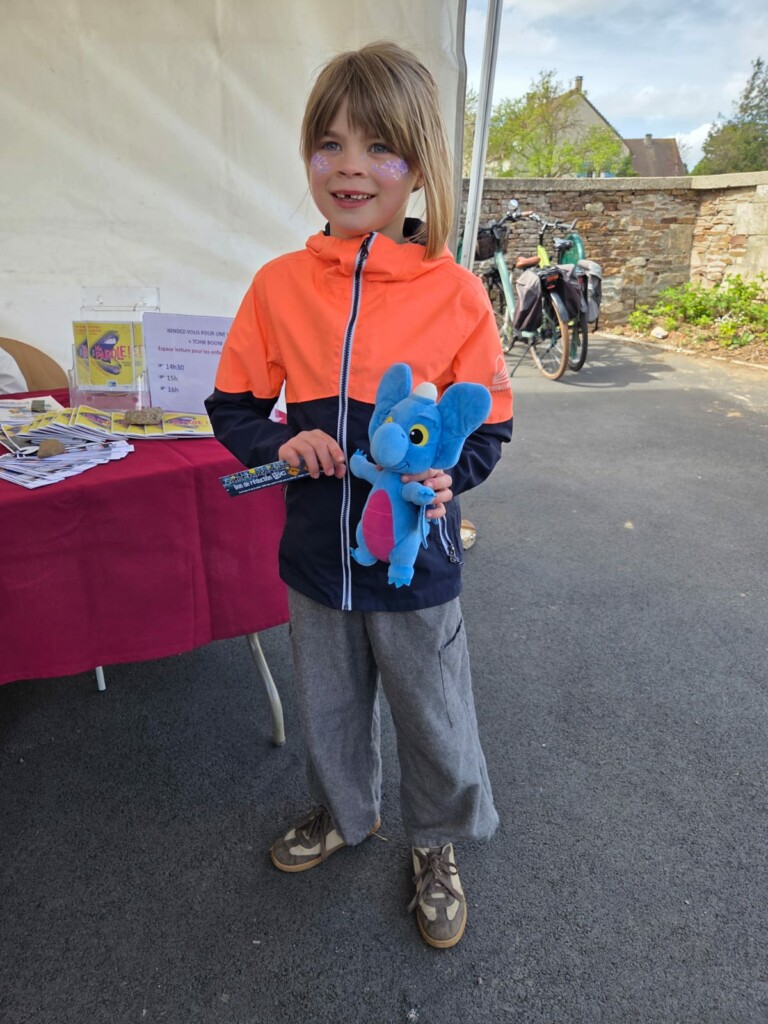 Enfant souriant, portant une veste orange, avec un jouet en peluche bleu, lors d'un événement à Feuguerolles-Bully.
