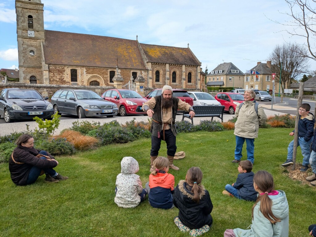 Un animateur en costume interagit avec des enfants assis sur l'herbe, devant une église à Feuguerolles-Bully.