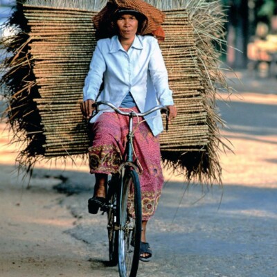 Femme à vélo transportant des matériaux naturels, sur une route rurale.